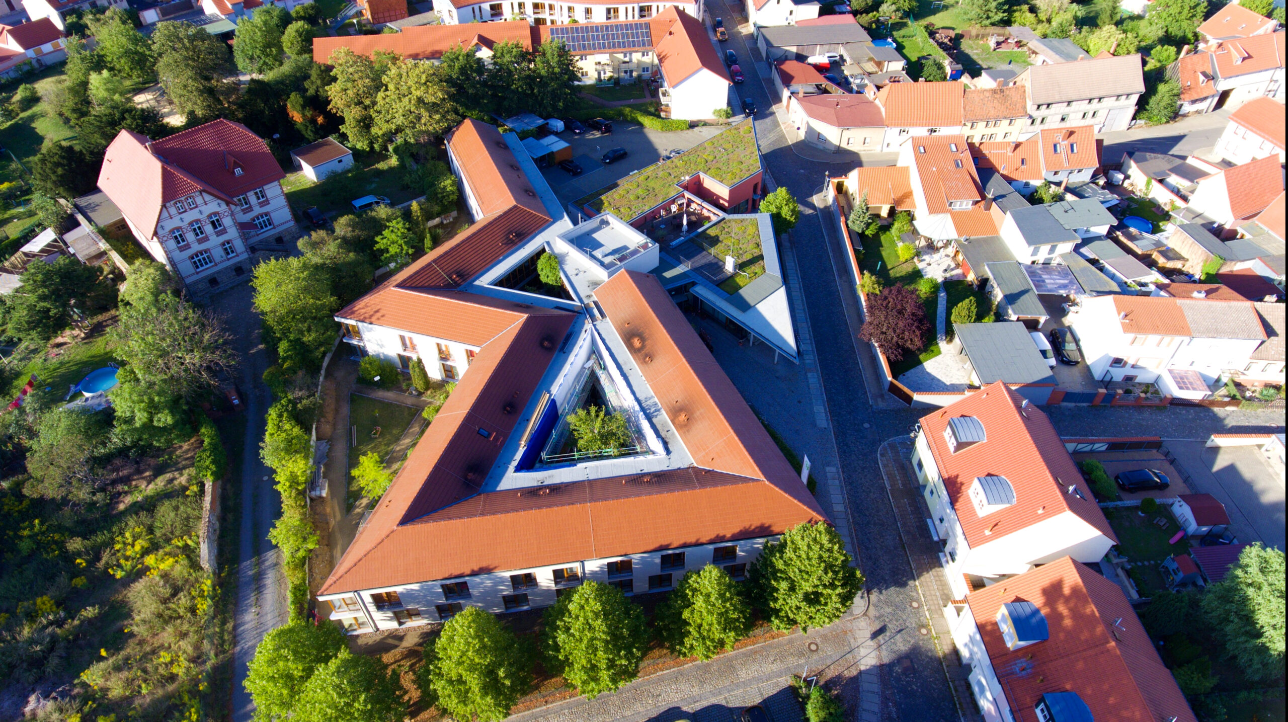 Luftaufnahme eines großen Gebäudekomplexes mit roten Dächern und dreieckigem Innenhof, umgeben von Wohnhäusern.