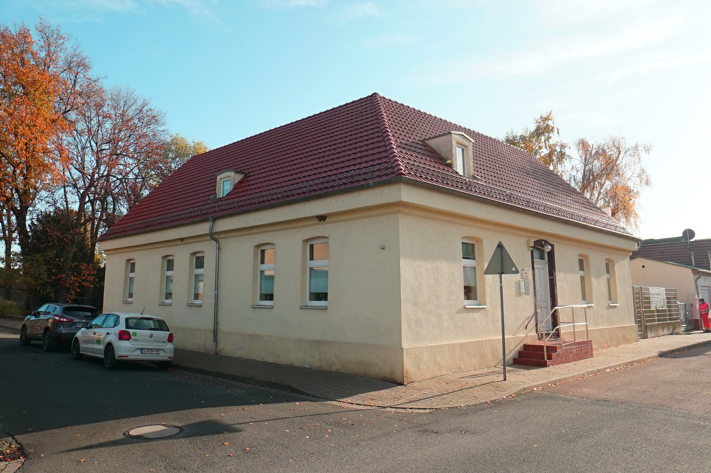 Helles Gebäude mit rotem Ziegeldach an einer Straßenecke; davor stehen geparkte Autos.