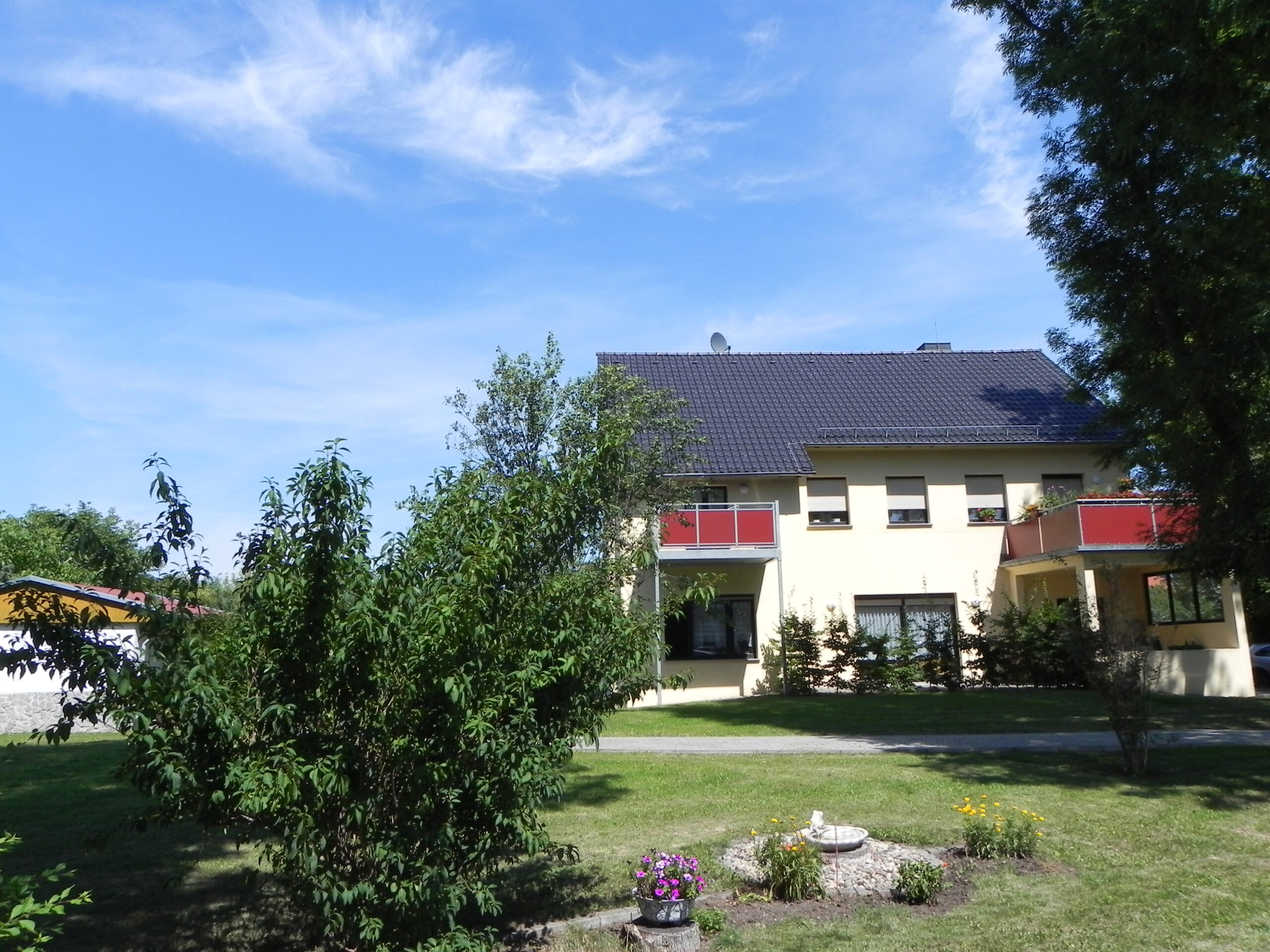 Gebäude mit dunklem Dach und roten Balkonen in einer grünen Gartenanlage unter blauem Himmel.