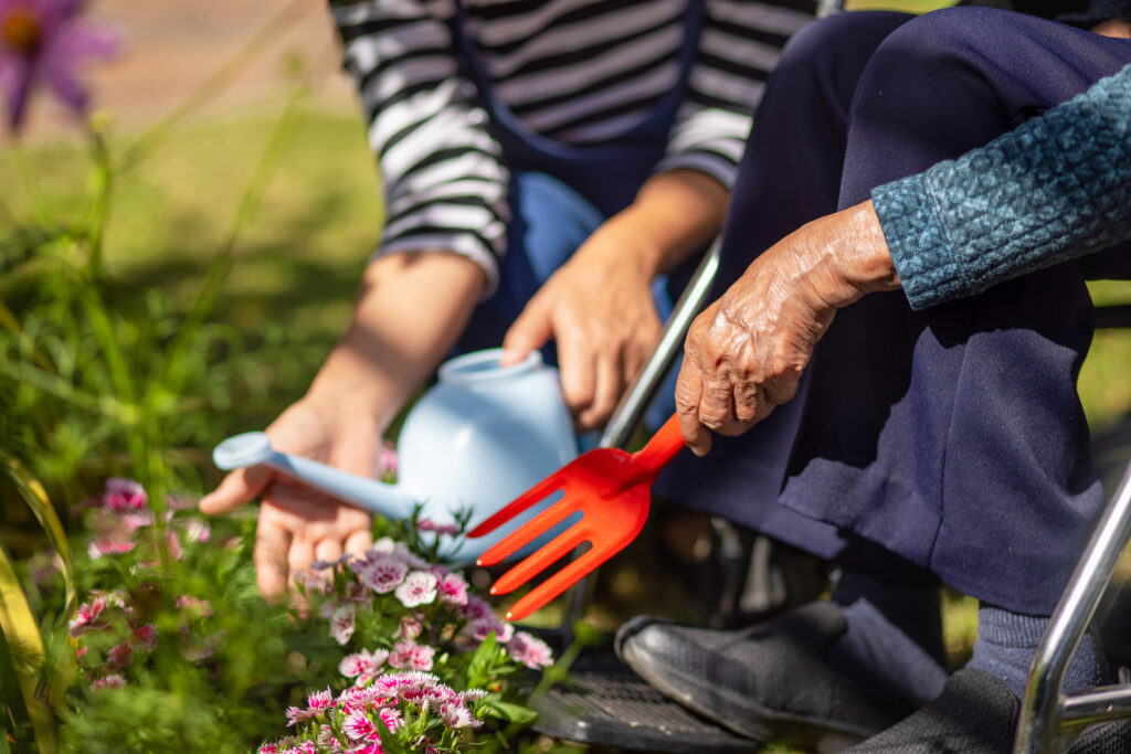 Zwei Personen gärtnern gemeinsam; eine Hand hält eine kleine Gießkanne, daneben ist eine rote Gartenschaufel an Blumen zu sehen.