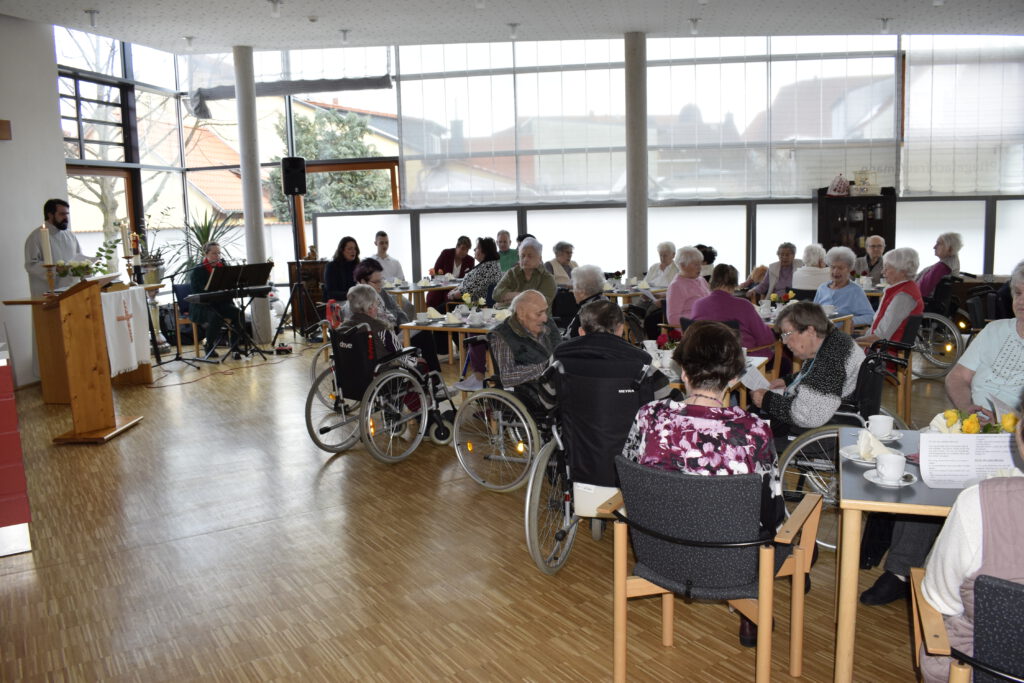 Bewohnerinnen und Bewohner sitzen an Tischen und singen gemeinsam mit dem Pfarrer Herrn Nürnberg