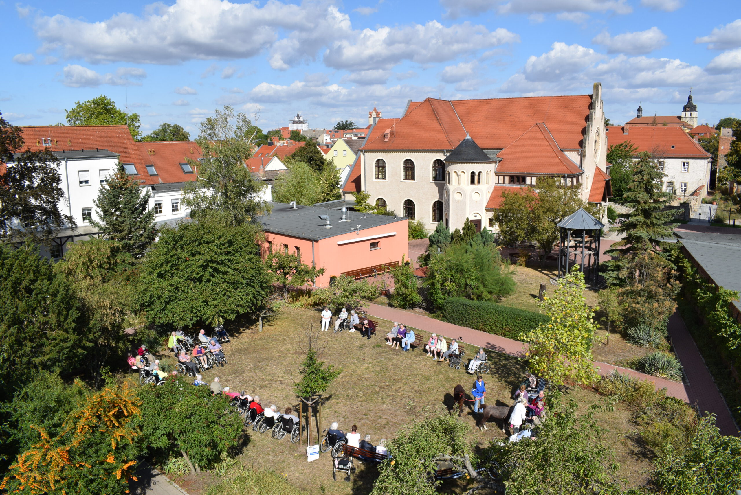 Blick von oben auf eine Anlage mit Garten und roten Dächern; viele Menschen sitzen draußen im Kreis.